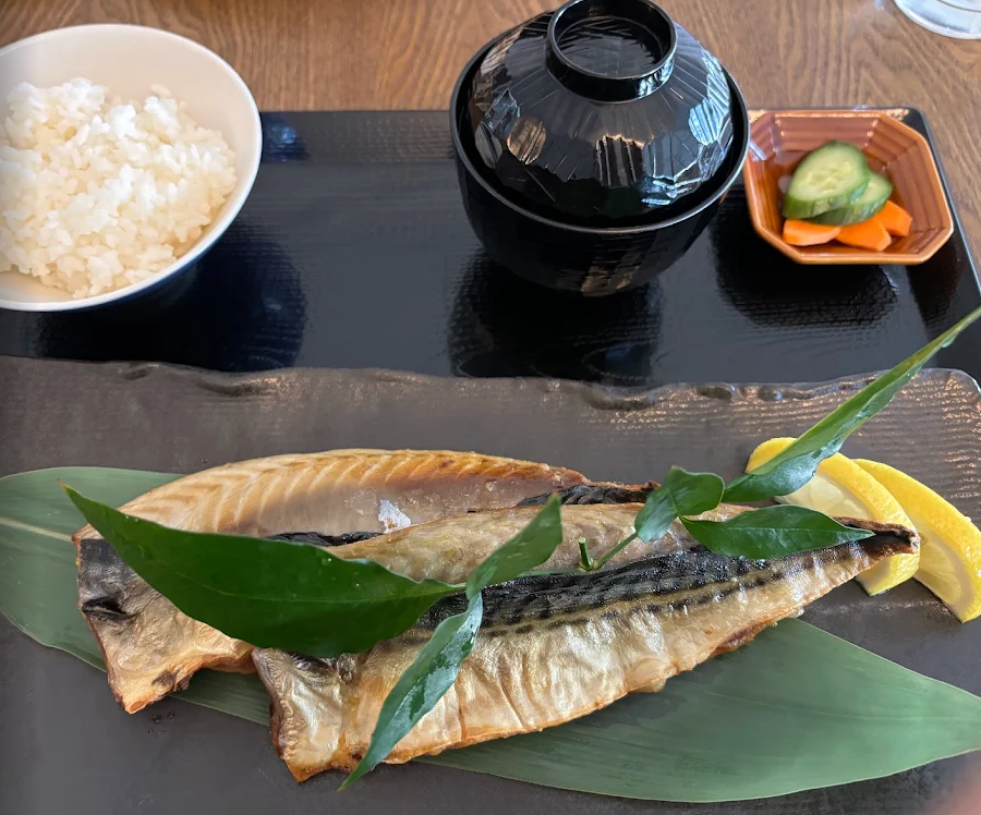 Plated grilled Hokke fish dish with green leaves on a dark surface, accompanied by a bowl of rice and a bottle.