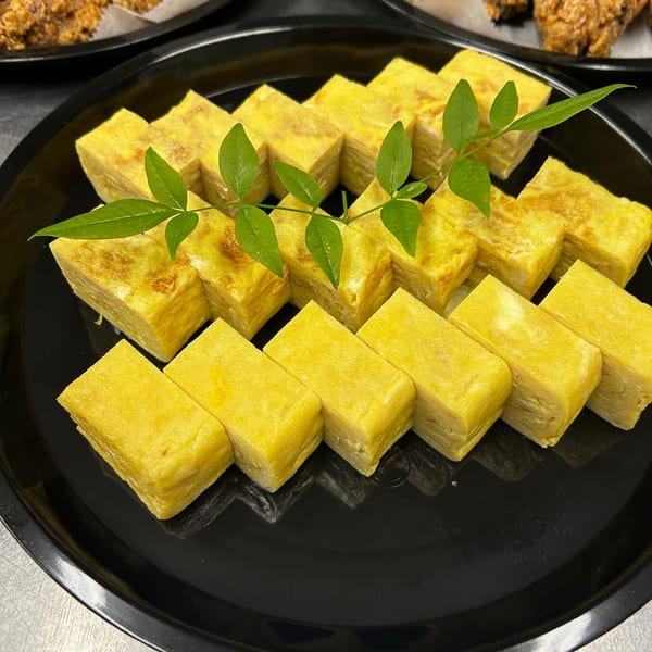 Rows of golden tamagoyaki (Japanese rolled omelet) garnished with green leaves on a black plate.